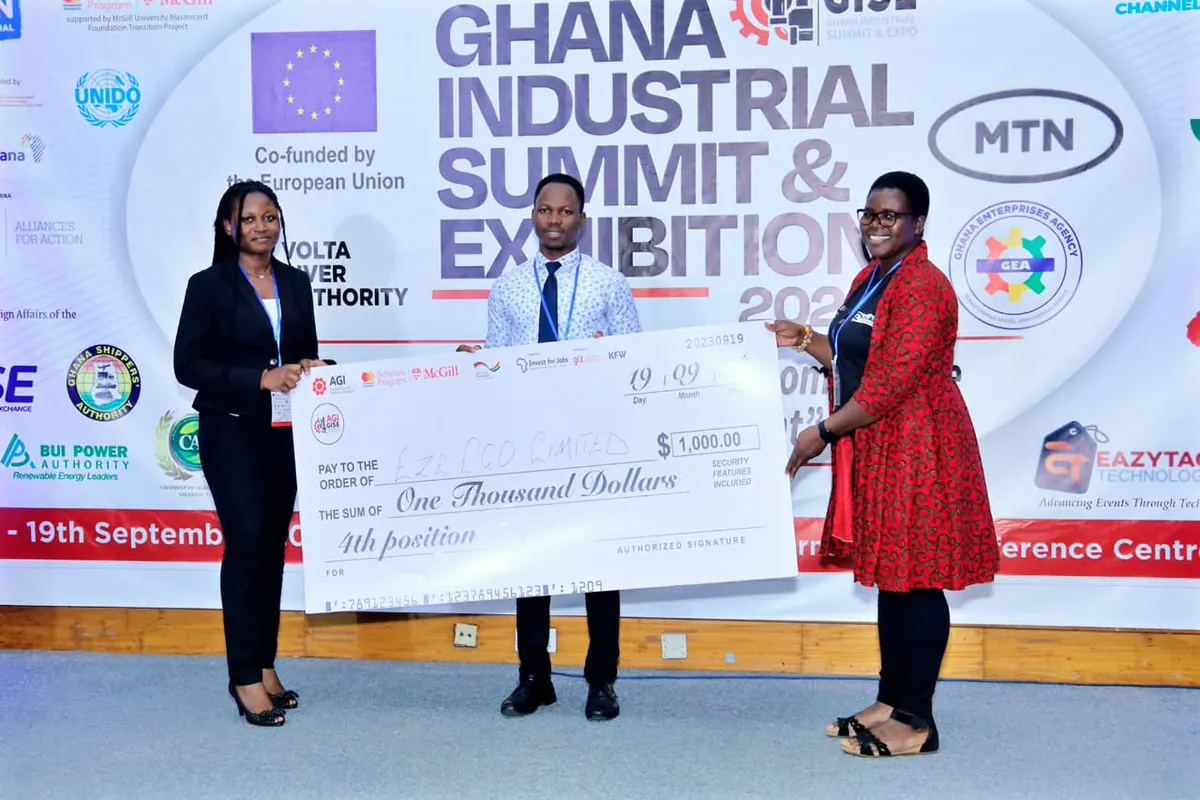 A team member receiving a 4th position cheque on stage at the Ghana Industrial Summit & Exhibition, between two presenters
