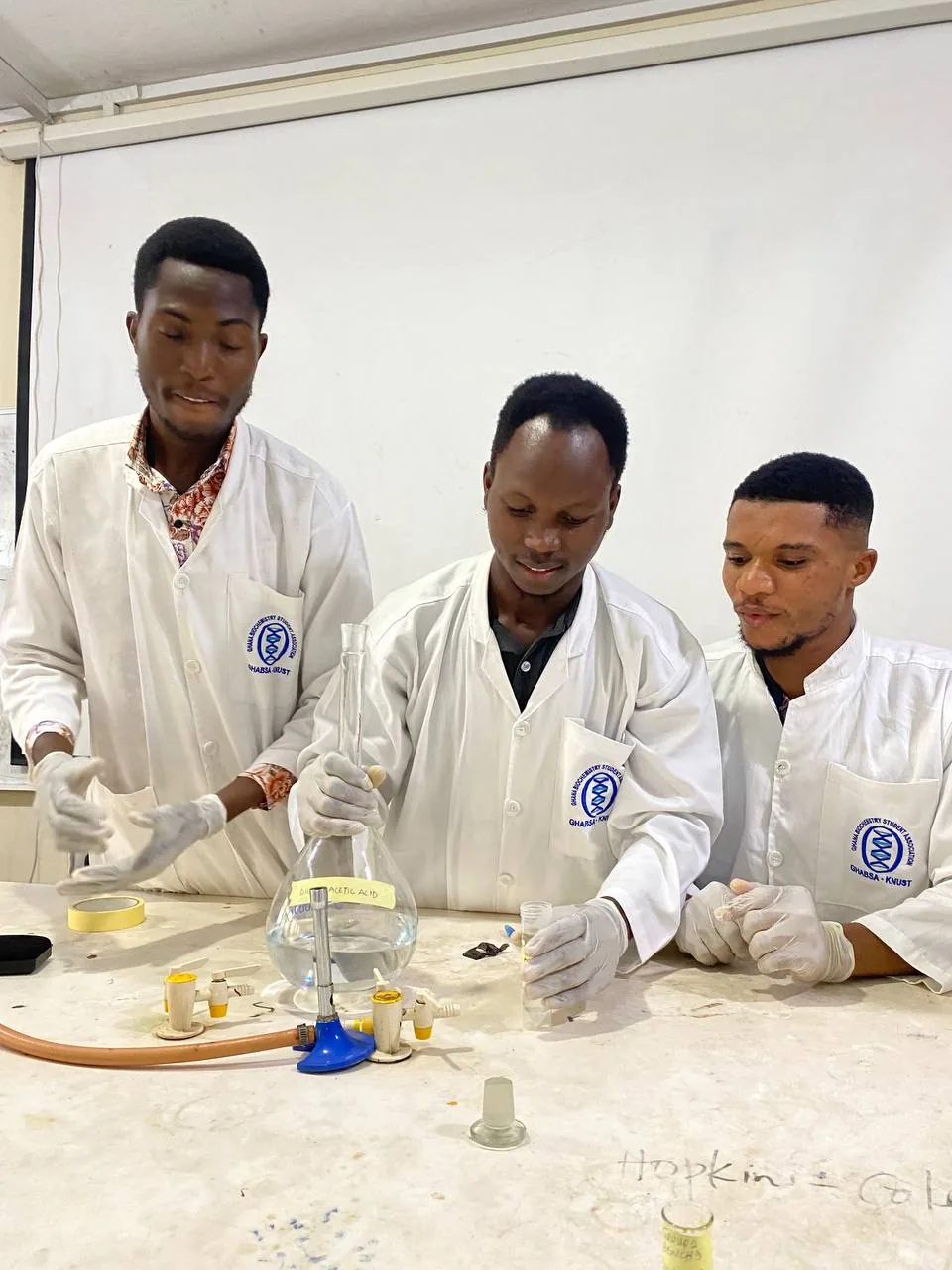 Three researchers in lab coats and gloves working with a volumetric flask and glassware at a laboratory bench